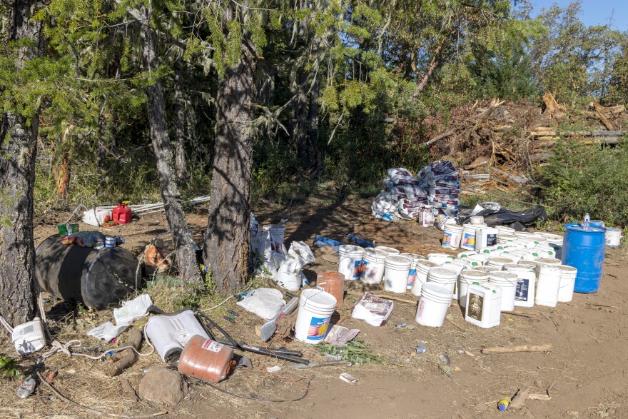 A large number of plastic containers and bags sit in a pile outside underneath a tree. Various other debris and litter is strewn about. The containers appear to have been used to store chemicals.